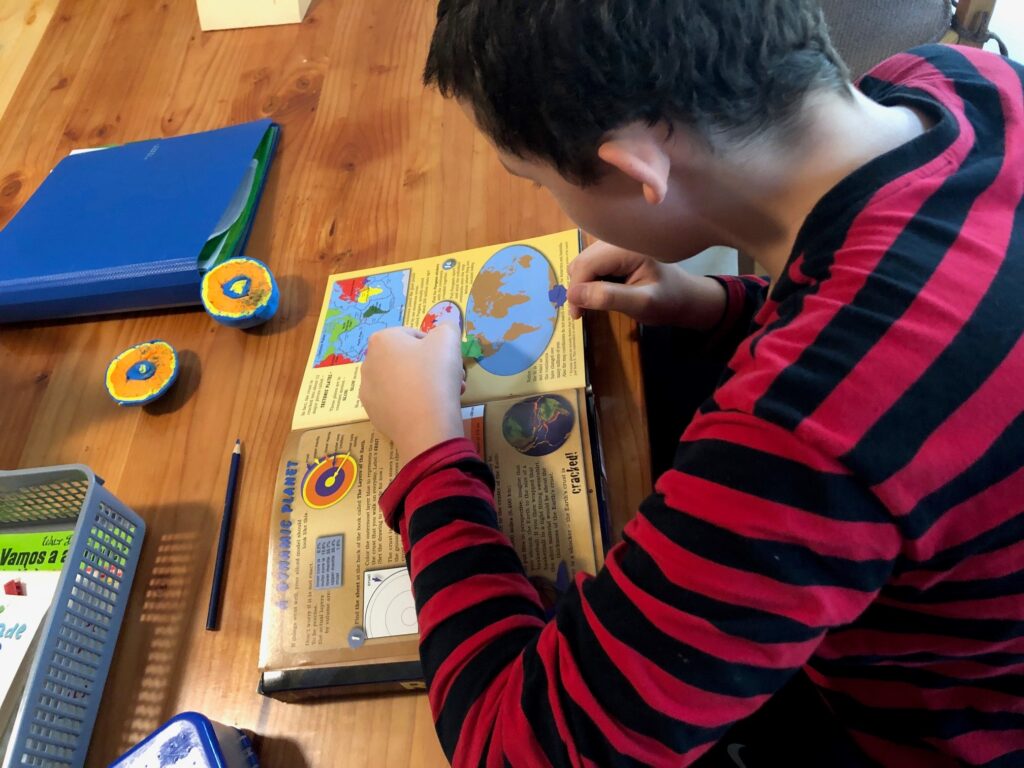 A teen boy focusing on a geology book, close by is a cut open model of the earth he made