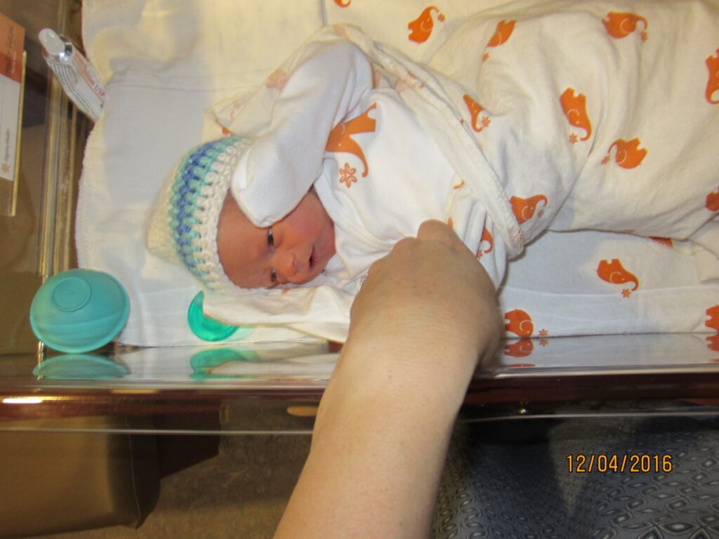 A swaddled baby in a hospital baby bed, his mother's hand holding his little hand