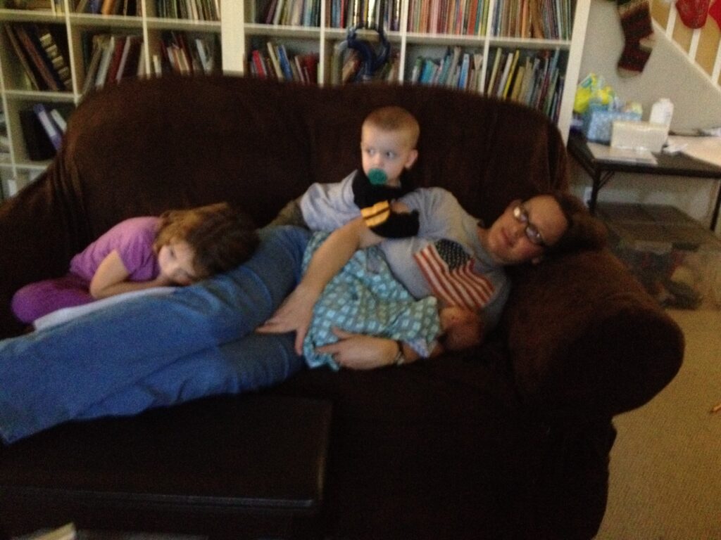 a woman lying on her side on a couch, a baby in her arm, a toddler on top of her, and a little girl resting on her leg
