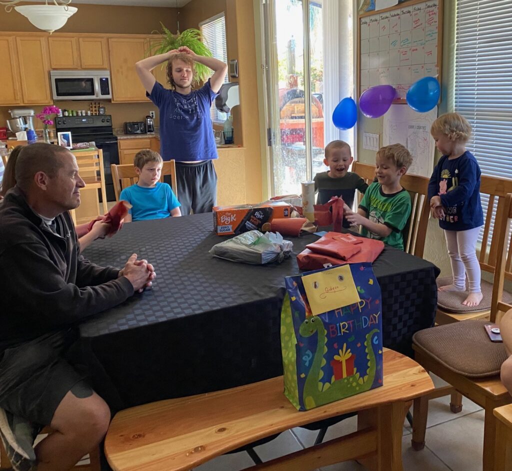 a family gathered at a table watching a boy open birthday presents