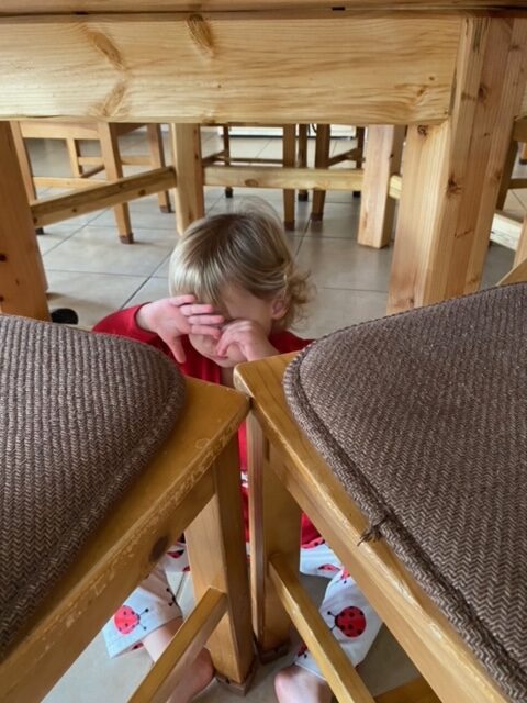 a toddler girl hiding under a table and behind chairs, her hands in front of her eyes and a frown