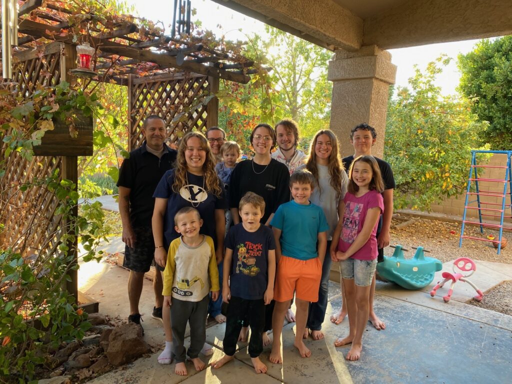 A family of a dad and mom and ten children standing on a back porch with a vine covered lattice and some trees