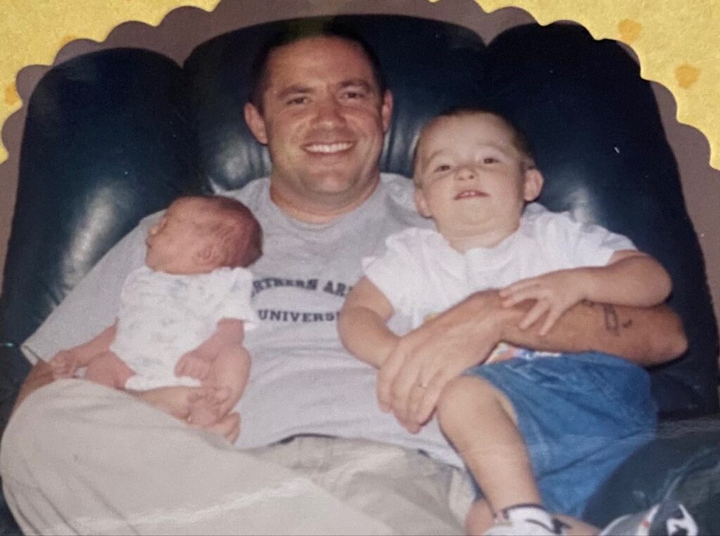 a smiling man in a recliner holding a baby girl and a toddler boy