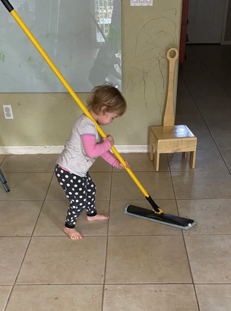 a toddler girl pushing a broom across a tile floor