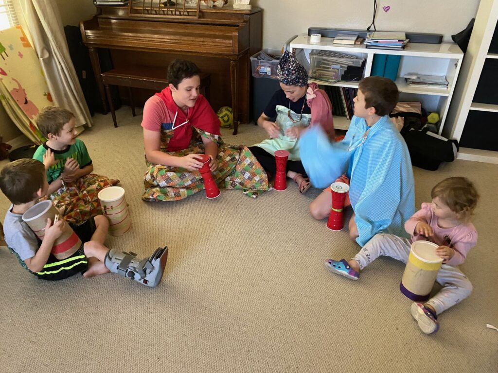 Children in a half circle, dressed in colorful fabric, beating homemade drums