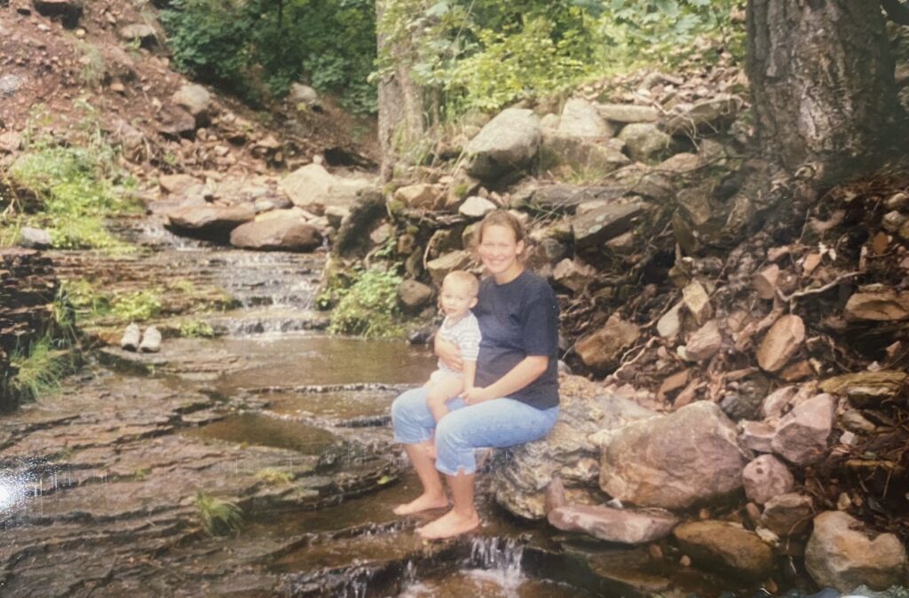 A pregnant woman sitting on a rock with her feet in a stream, a small boy on her lap